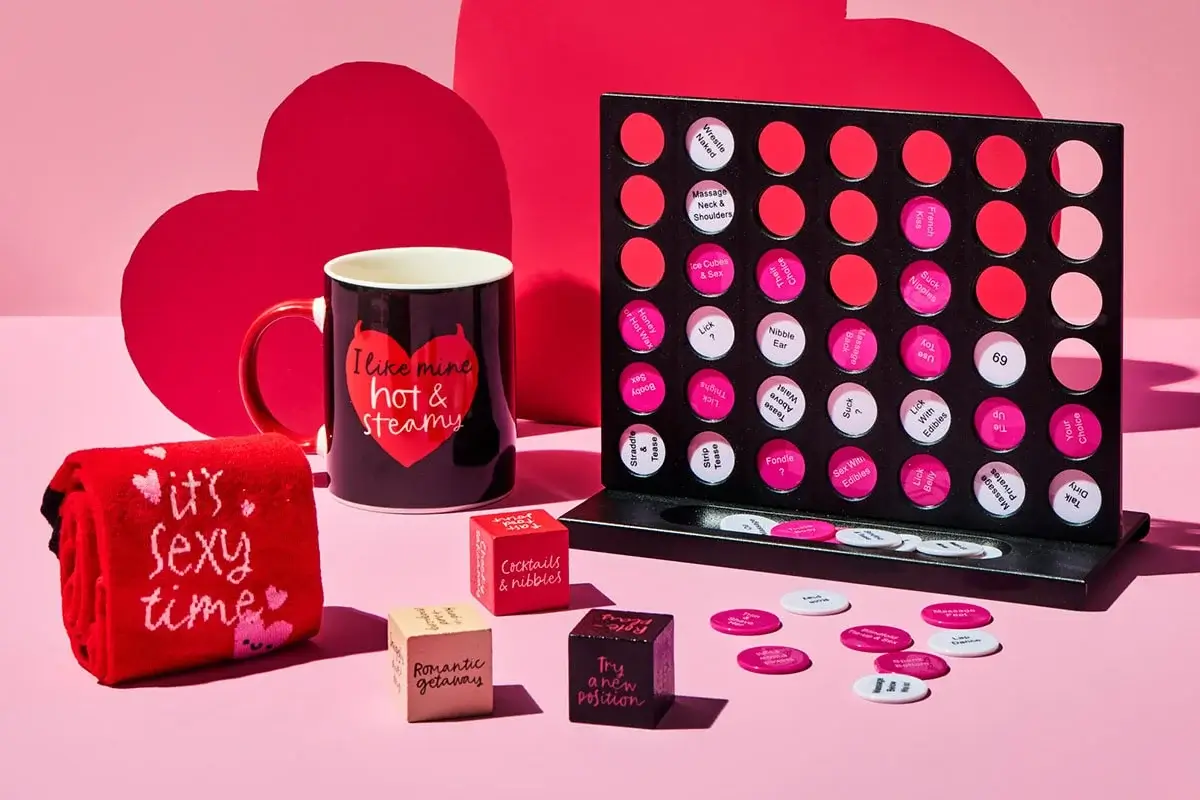 Valentine-themed gift set featuring a black mug with red text, a red towel with white text, a black and pink Connect Four-style game, and decorative heart-shaped props in the background.